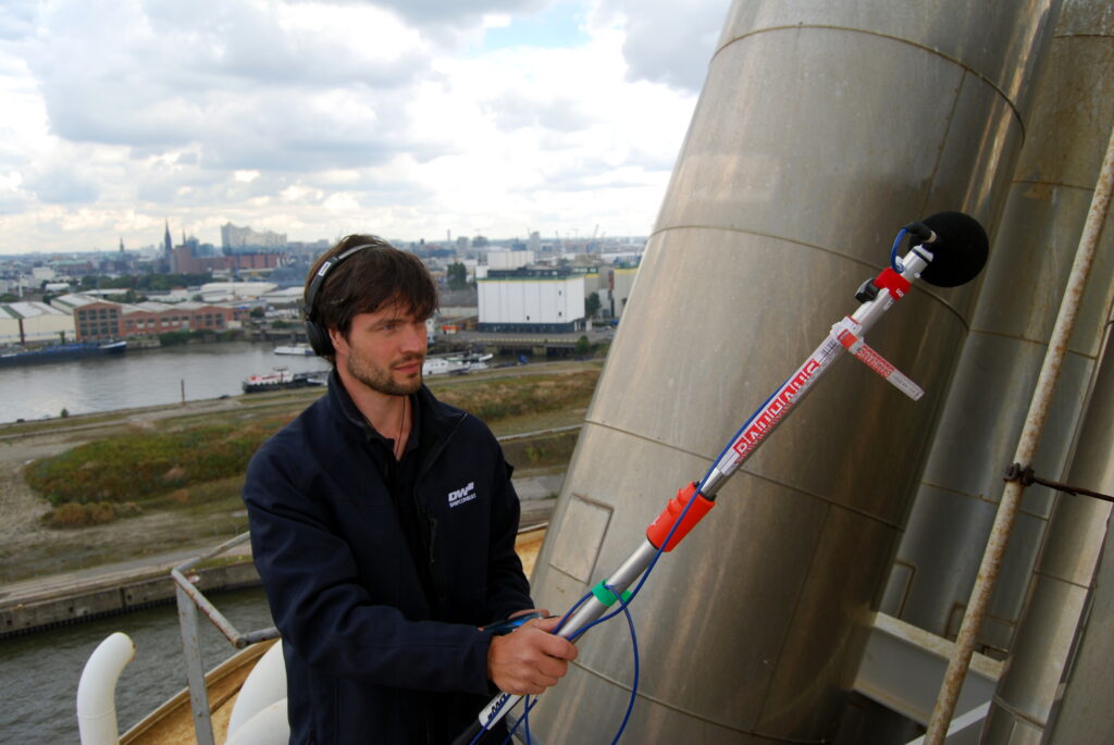Ein Schiffsakukstik Ingenieur steht in der Nähe eines Abgasaustritts eines Schiffes im Hamburger Hafen. Mit einem Luftschallmessgerät misst er den Schall im Abgas. 